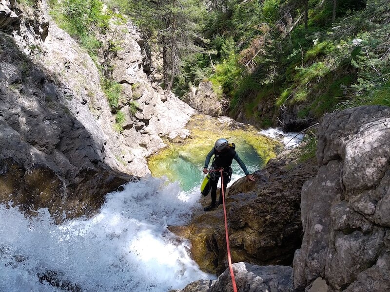 Personne équipée de cordes et casque progressant le long d’une cascade au cœur d’un canyon verdoyant.