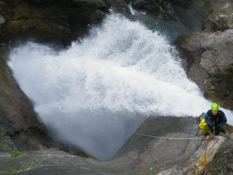 Cascade puissante projetant de l’eau en éventail, avec une personne en équipement de canyoning sur une paroi rocheuse.