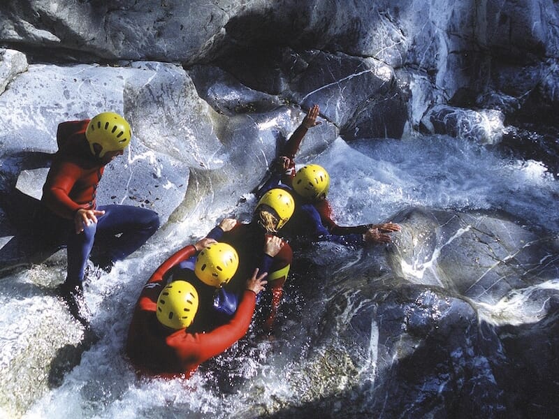 Groupe de six personnes en combinaisons et casques jaunes glissant ensemble dans un toboggan naturel rocheux.