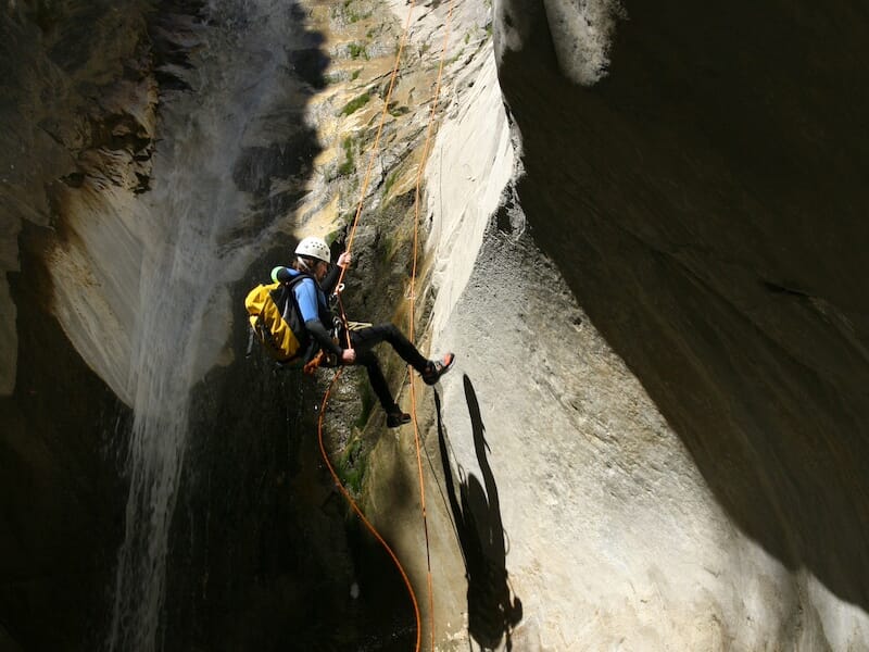 Pratiquant de canyoning suspendu par une corde le long d'une paroi humide, ombres marquées sur la roche éclairée.