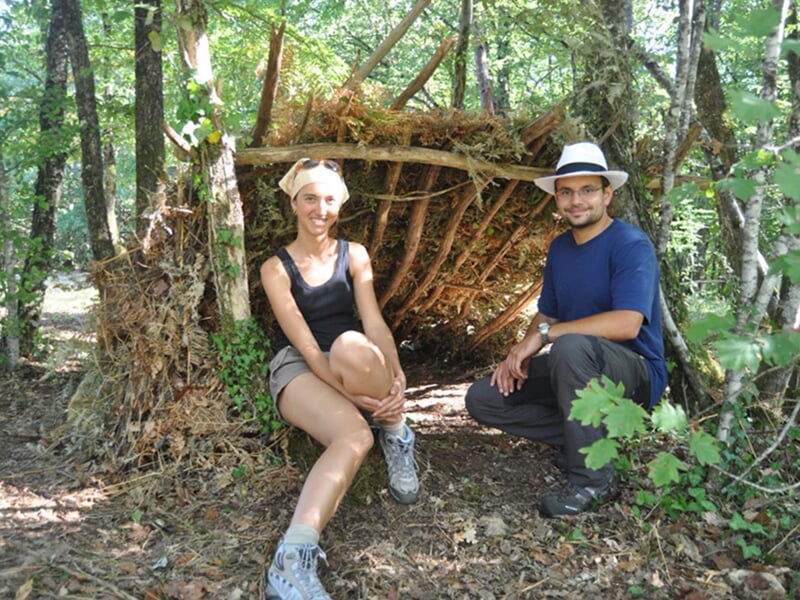 Deux personnes assises devant une cabane en bois faite de branches dans une forêt ensoleillée.