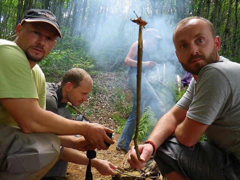 Trois hommes assis dans la forêt, concentrés sur un bâton tenu verticalement devant un feu de camp.