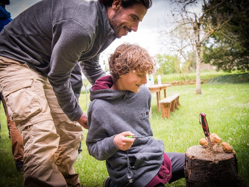 Groupe de jeunes assis sur l'herbe, souriants, dans un cadre naturel avec des arbres en arrière-plan.