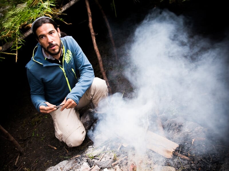 Homme agenouillé près d'un feu de camp dans une forêt, entouré de branches et de feuillage vert lors d'un stage de survie.