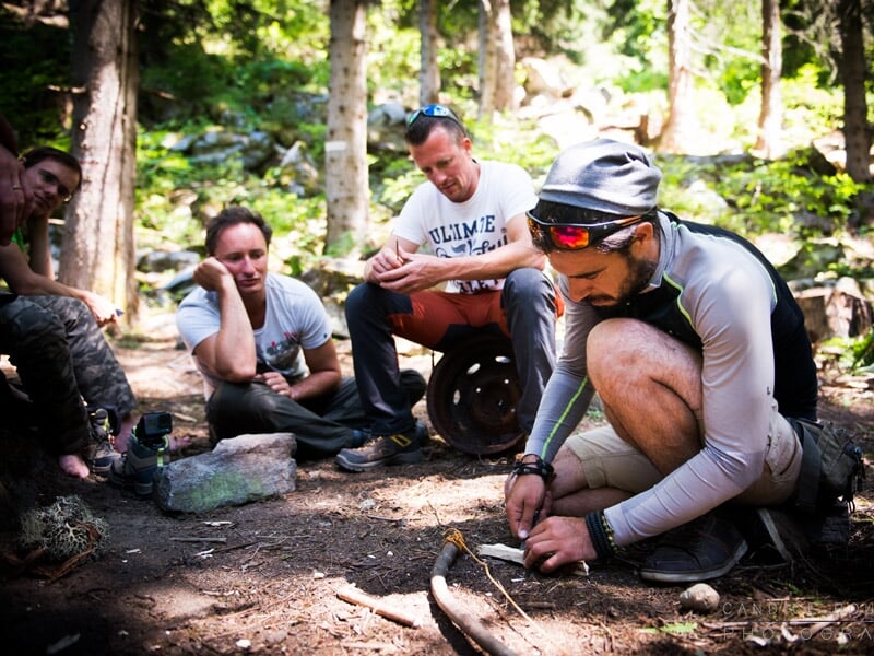 Un groupe d'hommes assis et agenouillé dans une forêt, l'un d'eux concentré sur l'affûtage d'un outil en bois lors d'un stage de survie