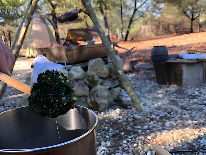 Une cuillère en bois tenant une boule de légumes verts au-dessus d'un pot en métal dans un cadre de camping en plein air avec une tente et un feu de camp flou en arrière-plan