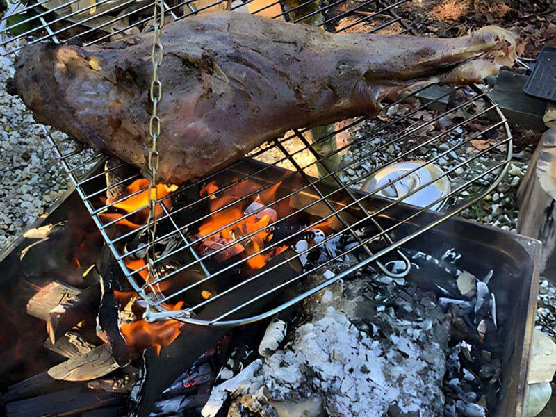 Un gigot d'agneau suspendu au-dessus d'un feu de camp, en train de rôtir sur une grille, avec des flammes visibles et des braises ardentes en dessous