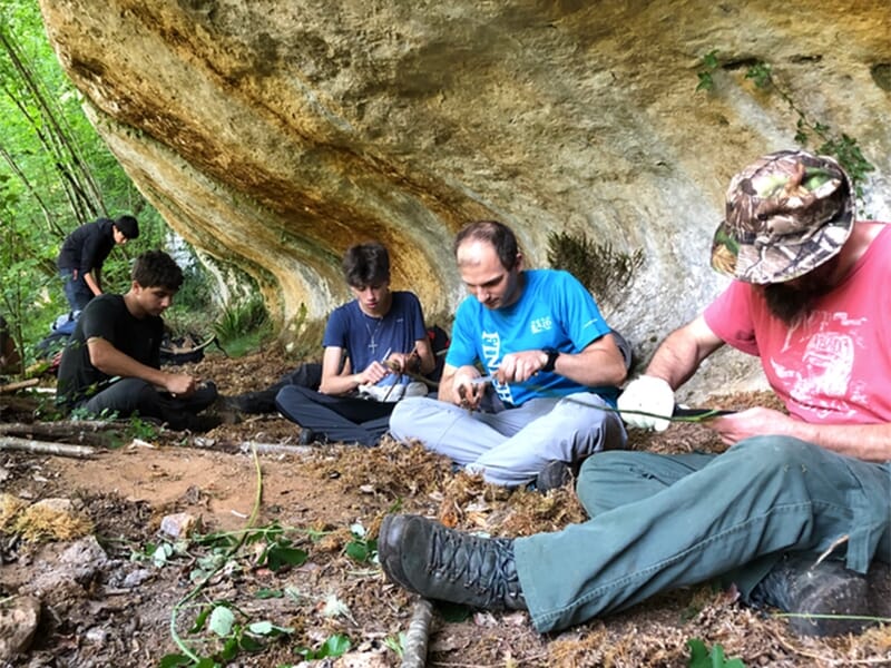 Un groupe de personnes assises sous une paroi rocheuse dans une forêt, en train de travailler le bois ou fabriquer des outils primitifs.