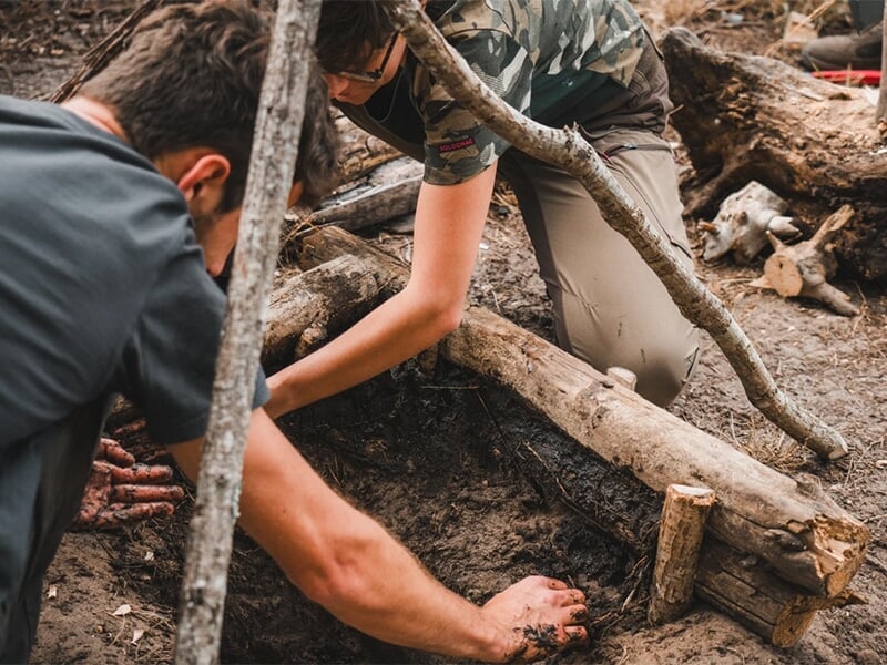 Deux personnes en train de construire une structure en bois, creusant dans le sol pour installer des branches sur un terrain de terre battue.