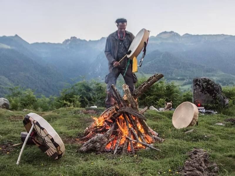 Homme debout près d'un feu de camp en montagne, jouant d'un tambour chamanique au coucher du soleil, lors d'un stage de survie.