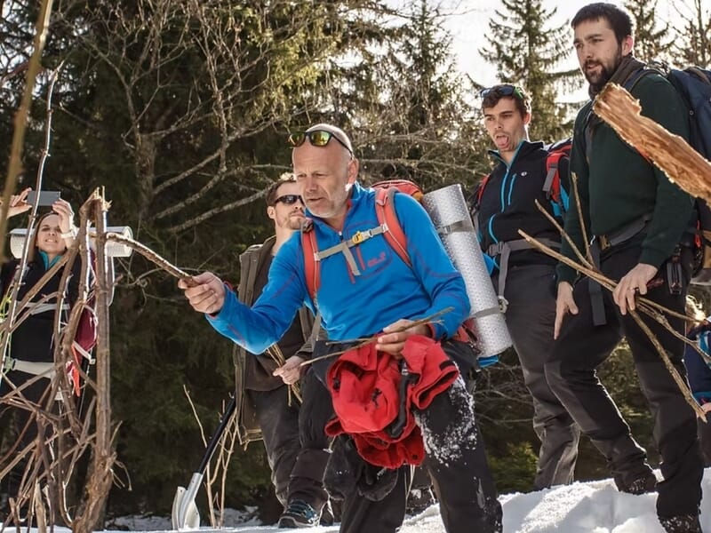 Homme en tenue de randonnée montrant un bâton en bois à un groupe lors d'un stage de survie en hiver dans le Haut-Jura.
