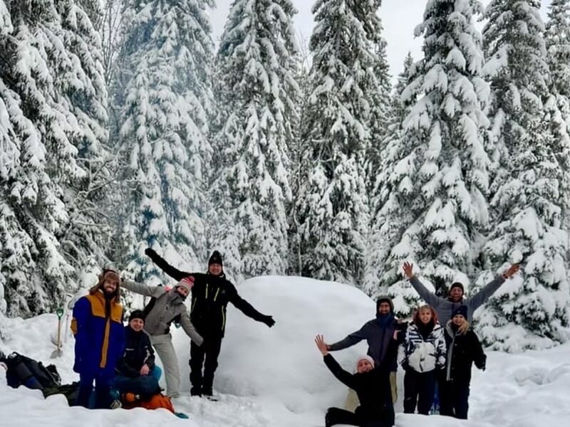 Groupe de neuf personnes en vêtements d'hiver posant joyeusement devant un igloo lors d'un stage de survie en hiver dans le Haut-Jura.