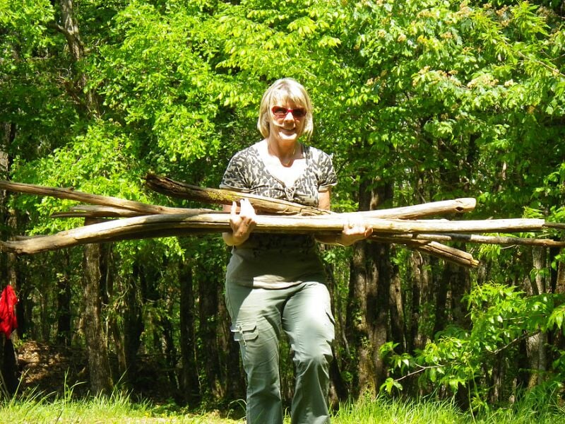 Femme souriante portant plusieurs branches de bois dans une forêt verdoyante en plein jour.