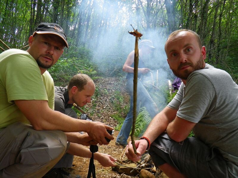 Groupe d'hommes assis dans la forêt, concentrés sur un bâton tenu verticalement devant un feu de camp.