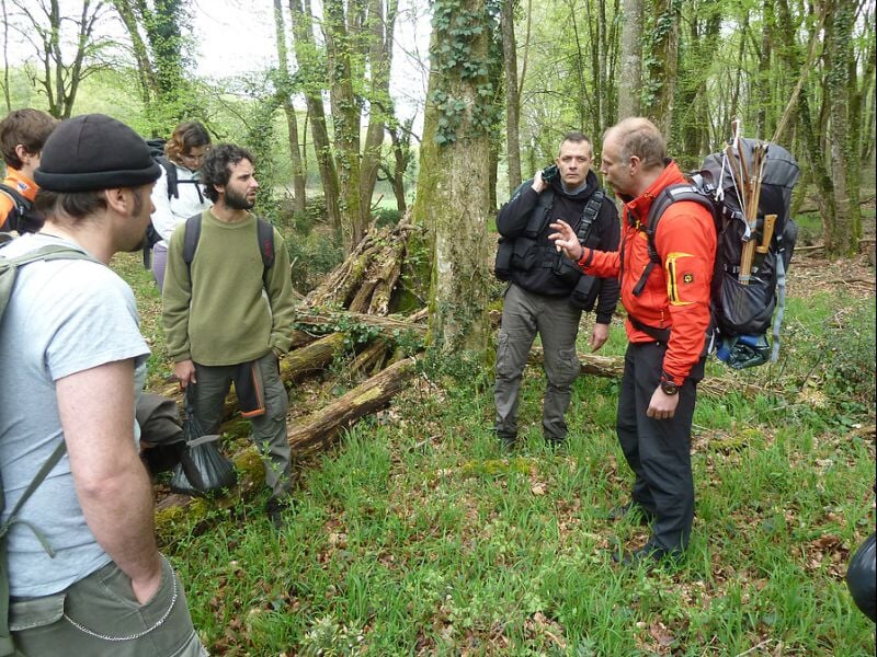 Homme en tenue rouge expliquant quelque chose à un groupe d'hommes en forêt, tous équipés de sacs à dos.