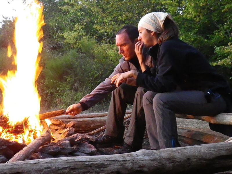 Deux personnes assises près d'un feu de camp en forêt, observant attentivement les flammes.