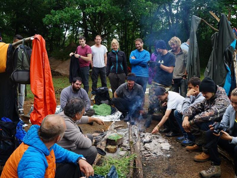 Groupe de personnes assises autour d'un feu de camp en forêt, discutant et partageant un moment convivial.