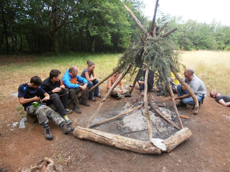 Personnes assises en cercle autour d'un feu de camp, avec une structure en bois au centre dans un espace dégagé.