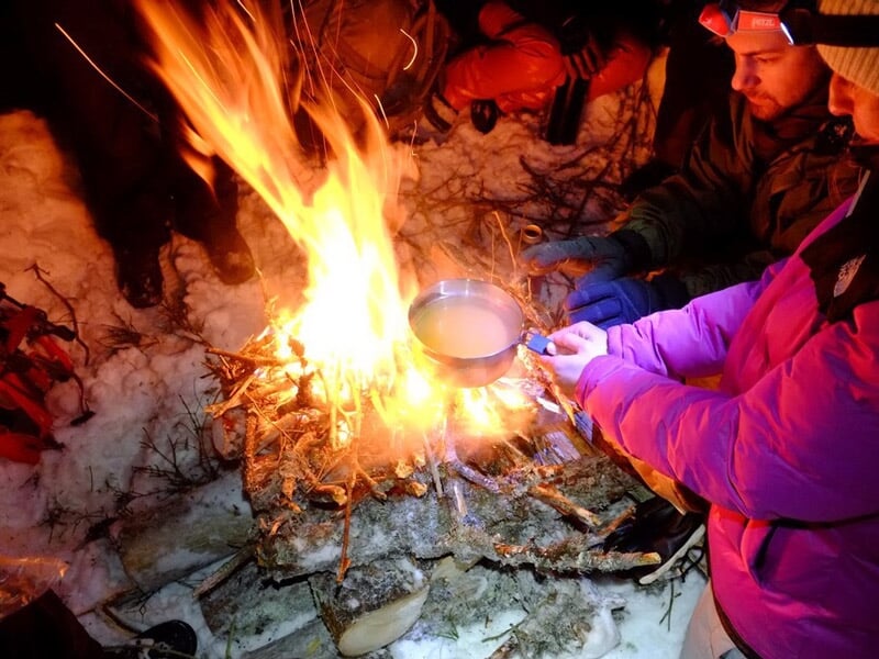 Groupe de personnes assises près d'un feu de camp allumé, une personne tient une tasse pour chauffer une boisson.