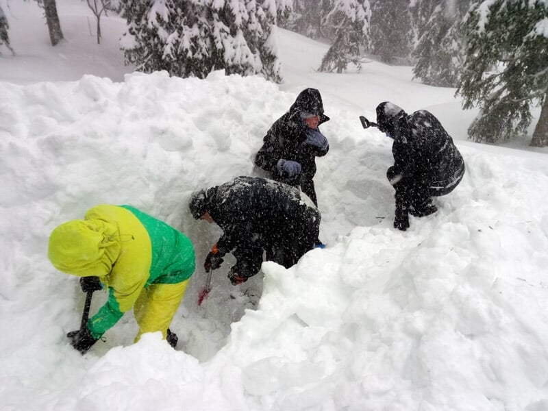 Quatre personnes creusent dans la neige épaisse sous une forte chute de neige, vêtues de vêtements d'hiver.
