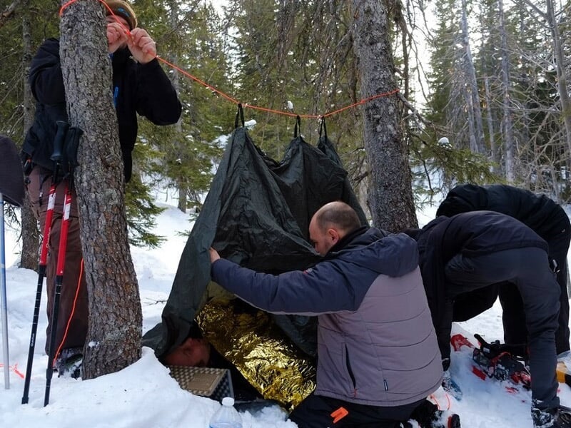 Trois personnes installent un abri de fortune en bâche noire entre deux arbres dans une forêt enneigée.