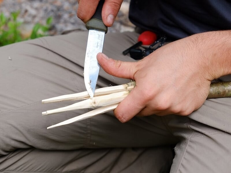 Une main taille des branches en pointes avec un couteau pour préparer du matériel de survie en forêt.