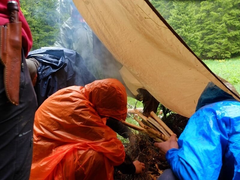 Deux personnes sous une bâche protègent un feu de camp allumé dans un environnement humide et boisé.