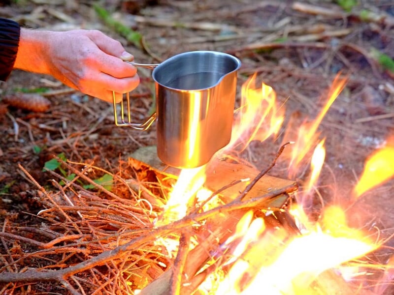 Une main tient une tasse métallique au-dessus d'un feu de camp allumé avec des brindilles sèches.