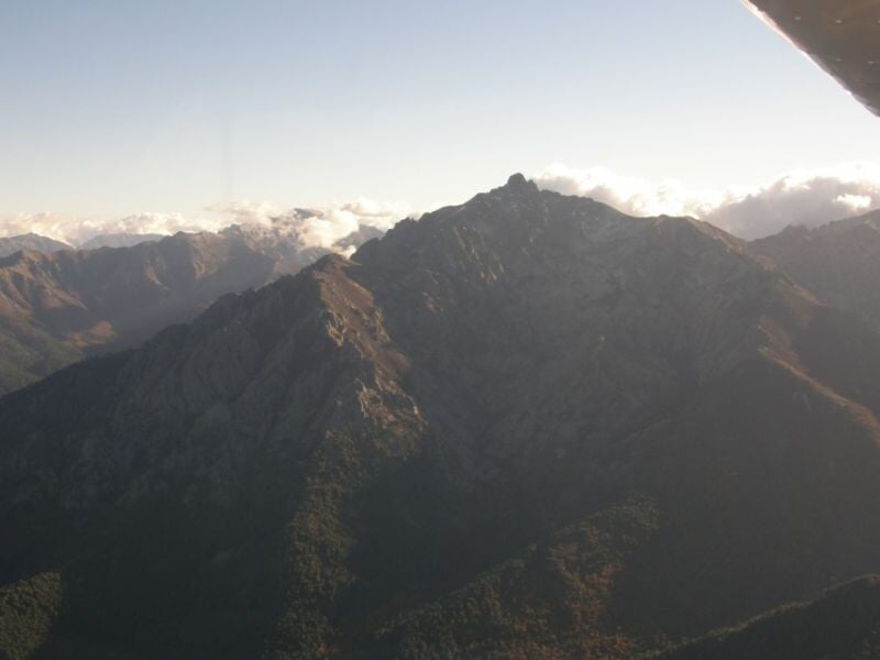 Vue aérienne d'une chaîne de montagnes sous un ciel clair avec des sommets rocheux et des vallées boisées.