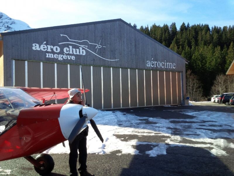 Homme debout près d'un petit avion rouge devant un hangar de l'aéro club de Megève par une journée ensoleillée.