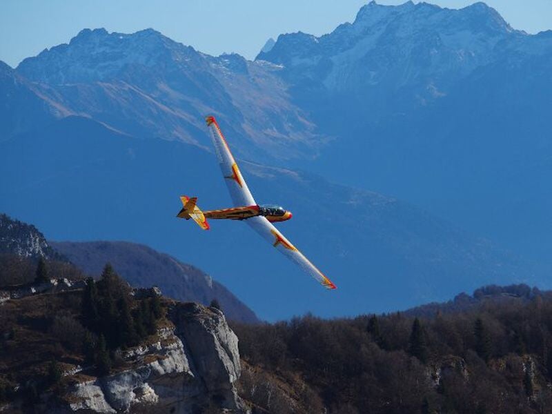 Planeur jaune et rouge en vol au-dessus d'une forêt avec des montagnes en arrière-plan sous un ciel bleu clair.