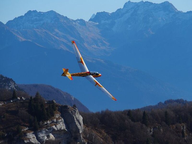 Planeur jaune et orange effectuant une manœuvre au-dessus d’un paysage montagneux, avec des sommets enneigés en arrière-plan. Vol libre dans un cadre alpin spectaculaire.