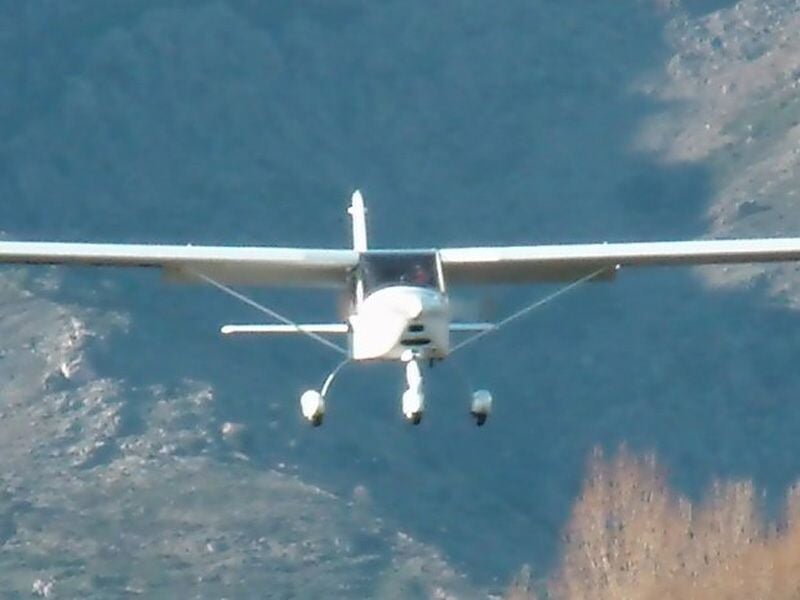 Avion léger en vol face à l’objectif, avec un arrière-plan montagneux. L’appareil à ailes hautes et train d’atterrissage fixe semble en approche ou en phase de vol stabilisé.