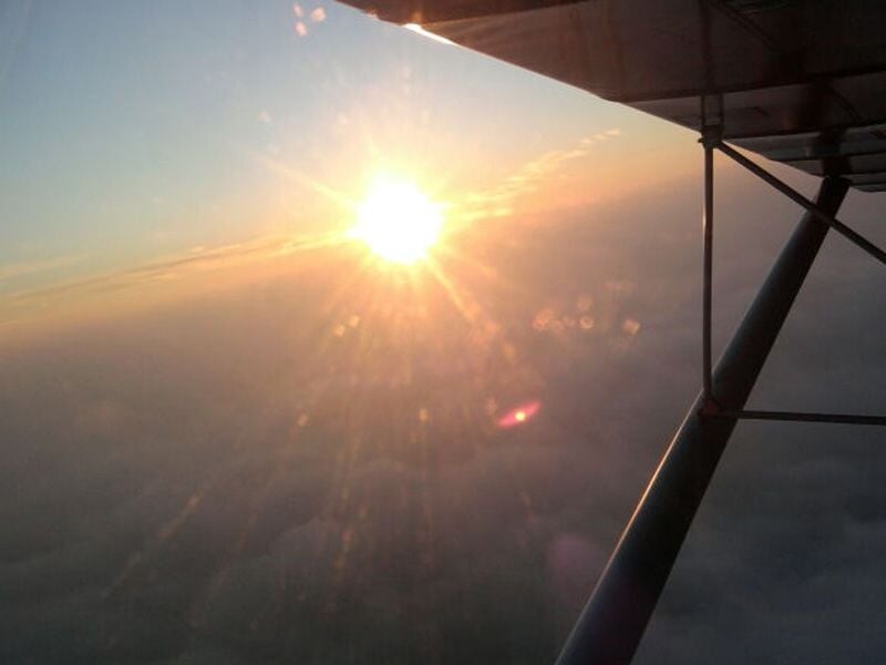 Coucher de soleil vu depuis un avion en vol, avec les rayons illuminant un ciel dégagé au-dessus d’une mer de nuages. L’aile et la structure de l’appareil sont visibles au premier plan.