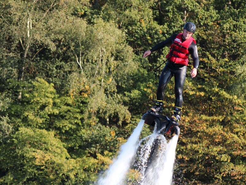 Personne en combinaison noire et gilet rouge pratiquant le flyboard au-dessus de l'eau avec un fond d'arbres verts.