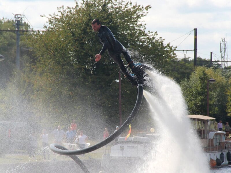 Homme en combinaison noire en train de voler avec un flyboard au-dessus de l'eau, entouré de spectateurs et d'arbres.