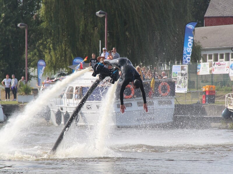 Flyboarder effectuant une figure acrobatique au-dessus de l'eau avec un bateau et des spectateurs en arrière-plan.