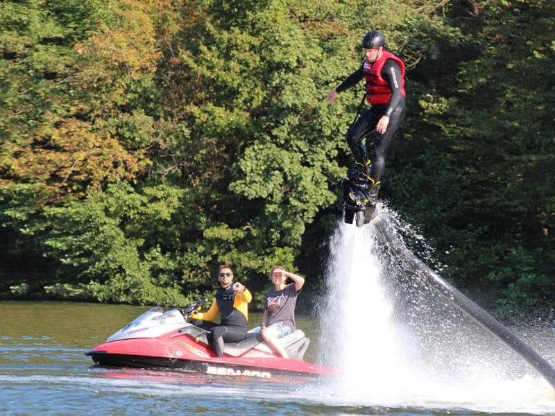 Personne en combinaison noire et gilet rouge sur un flyboard propulsé par de l'eau, avec deux personnes sur un jet-ski à côté.