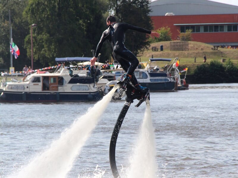 Homme en combinaison noire réalisant un saut en flyboard sur une rivière avec des bateaux et un bâtiment en arrière-plan.