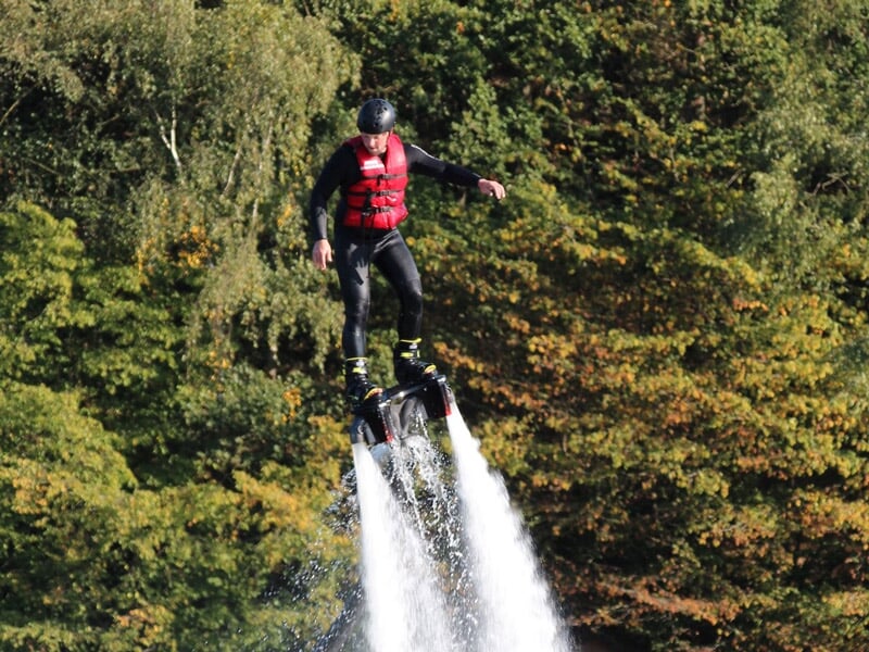 Flyboarder en combinaison noire et gilet rouge s'élevant au-dessus de l'eau avec un fond d'arbres verts et ciel bleu.