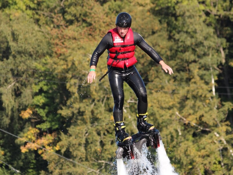 Personne en combinaison noire et gilet rouge en train de voler avec un flyboard au-dessus de l'eau, entourée d'arbres verts.