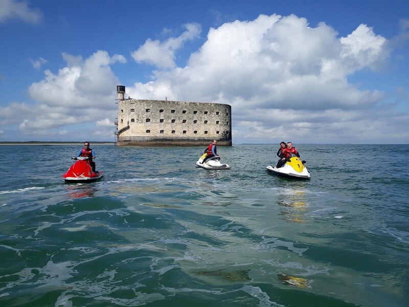 Trois personnes sur des jetskis rouges, blancs et jaunes naviguent près d'un fort en mer sous un ciel nuageux.