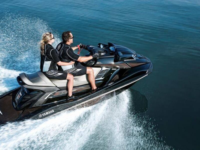 Deux personnes en combinaison noire pilotent un jetski Yamaha gris sur une mer calme et bleue.