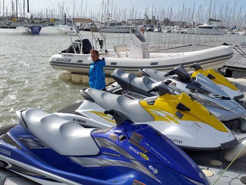 Plusieurs jetskis jaunes, blancs et bleus sont amarrés dans un port avec un homme en veste bleue.
