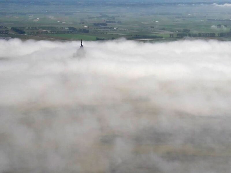 Vue aérienne du Mont Saint-Michel émergeant d'une mer de nuages avec paysage verdoyant en arrière-plan.
