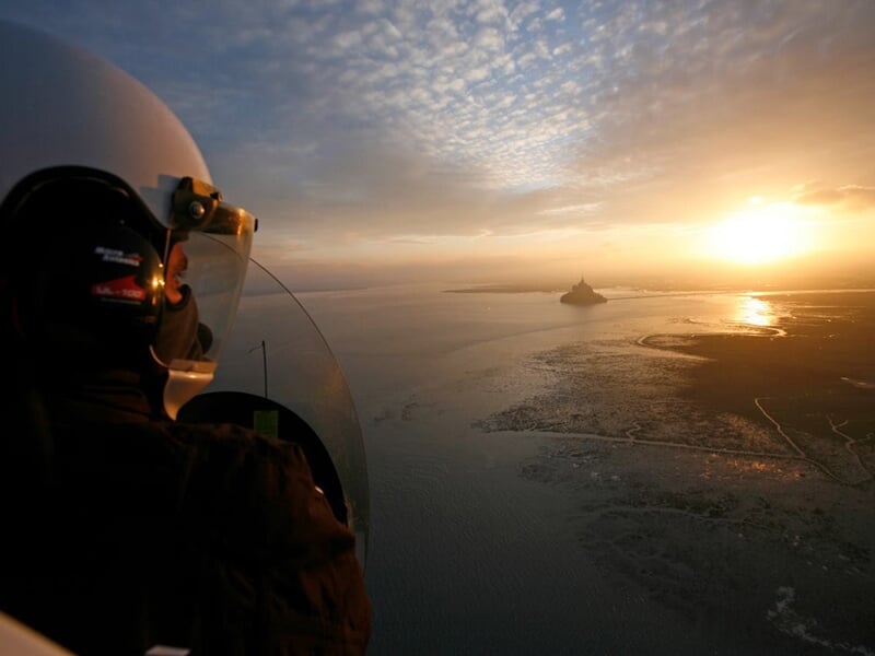 Pilote en casque dans un ULM survolant le Mont Saint-Michel au coucher du soleil avec ciel coloré.