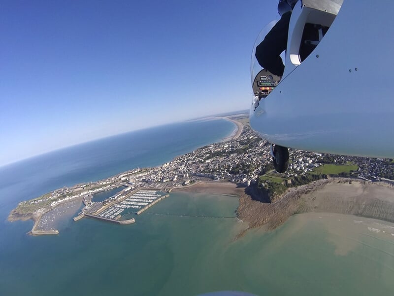 Vue depuis un ULM montrant une côte avec port, plage et mer sous un ciel bleu clair sans nuages.