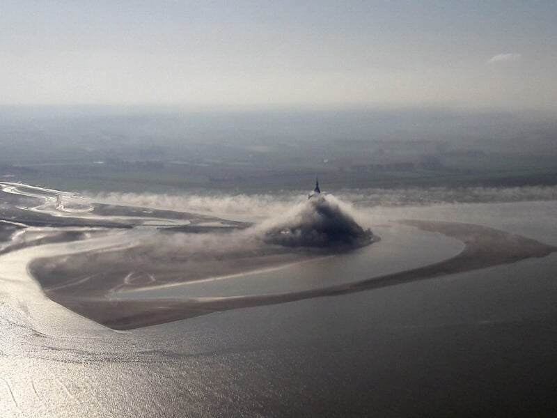 Mont Saint-Michel entouré de vasières et d'eau, vu depuis un ULM en vol avec lumière douce.