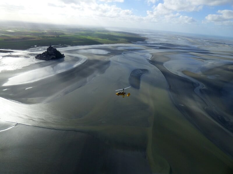 ULM jaune volant au-dessus des vasières et de l'eau près du Mont Saint-Michel sous un ciel clair.