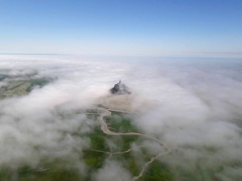 Mont Saint-Michel vu du ciel, entouré de nuages bas et de verdure, paysage naturel et paisible.
